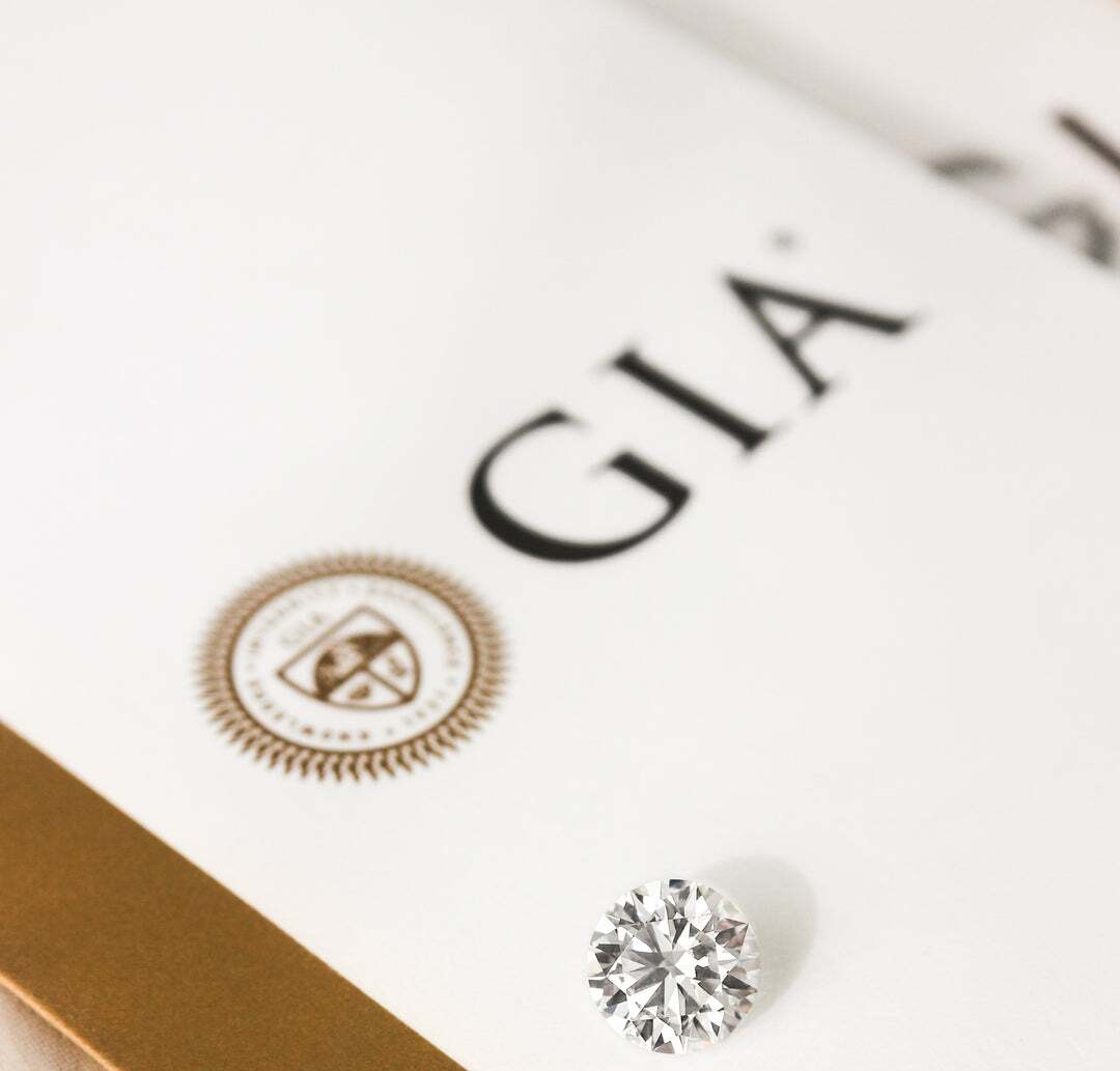 A round loose diamond is placed on top of a certificate from the Gemological Institute of America (GIA) for earth mined or natural diamonds. Behind it is a report from the International Gemological Institute (IGI).